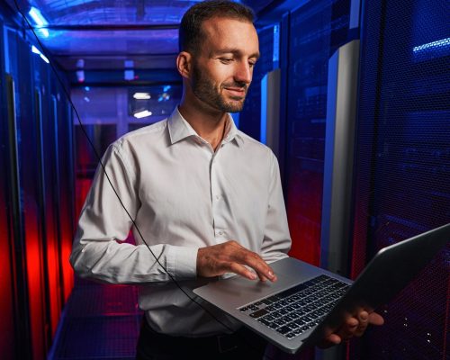 Caucasian programmer is looking to the screen of laptop while standing near the server rack